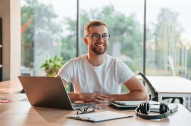 Oturuyor ve gülümsüyor. Modern ofiste resmi giyinmiş olumlu bir adam çalışıyor..