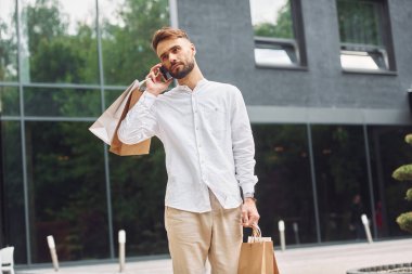 Büyük pencereli binaya karşı. Şık genç adamın alışveriş günü var. Çantalarla dışarıda..