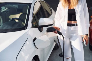 Otomobil şarj için bekliyor. Beyaz elbiseli genç bir kadın gündüz vakti elektrikli arabasıyla geliyor..
