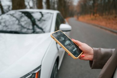 Elinde akıllı telefonla. Şarj işlemi. Kadın dışarıda, yolda elektrikli arabasının yanında duruyor..