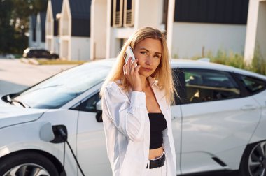 Telefona sarılıp onunla konuşmak. Beyaz elbiseli genç bir kadın gündüz vakti elektrikli arabasıyla geliyor..