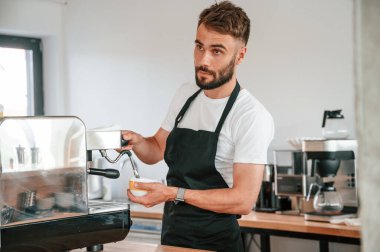 Ayakta ve kahve doldururken. Beyaz gömlekli ve siyah önlüklü kafe çalışanı içeride..