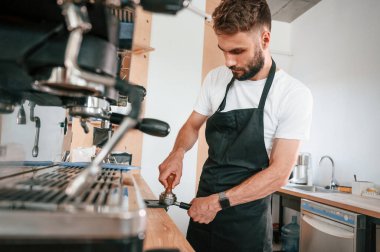 Kahve makinesini kullanarak. Beyaz gömlekli ve siyah önlüklü kafe çalışanı içeride..