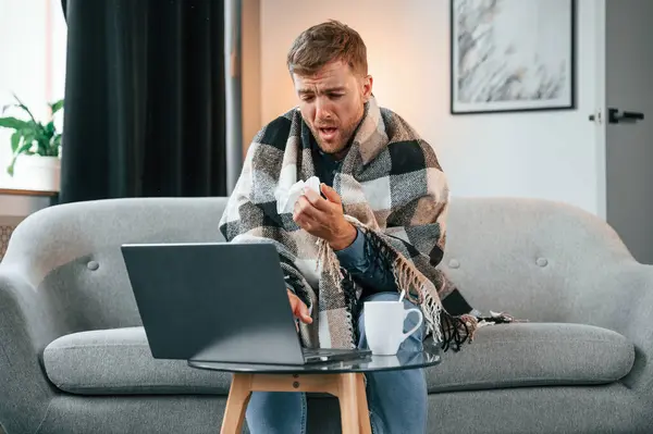 stock image Sitting and sneezes. Sick man is on the sofa at home by the laptop. Conception of illness.