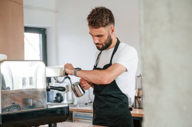 Ayakta ve kahve doldururken. Beyaz gömlekli ve siyah önlüklü kafe çalışanı içeride..
