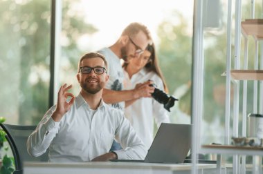 Kamerada bir kadına fotoğraf gösteren bir adam. Modern ofiste resmi kıyafetli üç kişi birlikte çalışıyor..