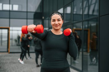 Gülümsüyor, dambıl kullanarak egzersiz yapıyor. Bir grup sportif kadın dışarıda, siyah binanın yakınında..