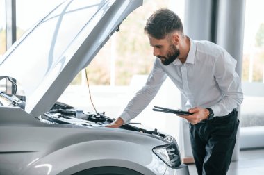 Arabanın kaputunun altına bakıyor. Beyaz elbiseli genç adam araba galerisinde..