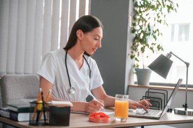 Dizüstü bilgisayarla çalışıyor. Elma suyu ve ölçü uzunluğu masanın üzerinde. Genç, güzel bayan doktor..