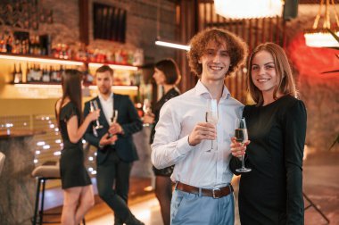 Kadın ve erkek ayakta duruyor ve poz veriyor. Güzel giyinmiş bir grup insan içeride yeni yılı kutluyorlar..