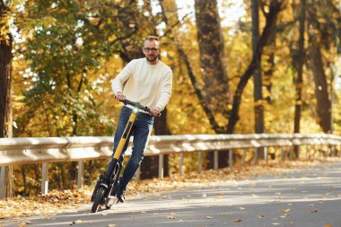 Gözlüklü ve rahat giyimli bir adam yolda elektrikli scooter kullanıyor..