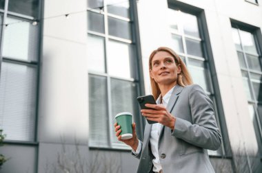 Bir bardak içki ve telefonla. Resmi giysili kadın iş binasının yakınında dışarıda..