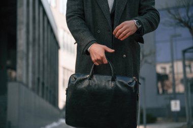 Siyah giysili adam dışarıda duruyor ve çantasını tutuyor..