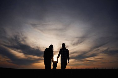 Baba, anne ve küçük bebeğin siluetleri gün batımına karşı kırda dramatik bir gökyüzü..