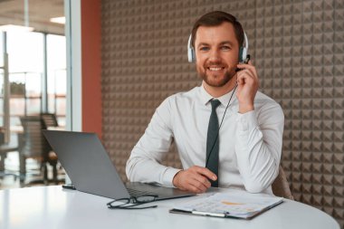 Kulaklıklardan. Resmi kıyafetli yakışıklı ofiste dizüstü bilgisayarla çalışıyor..