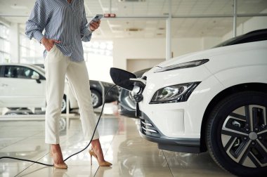 Ayakta duran, akıllı telefonu tutan, otomobil şarj oluyor. Elektrikli arabalı bir kadının yakın görüntüsü.