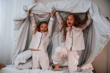 Yatak odasında, ellerinde battaniyeyle. İki küçük kız evcil odada oynuyorlar ve eğleniyorlar..