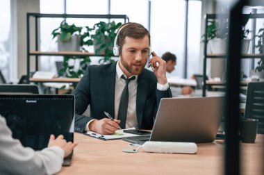 Kulaklıkla. Takım elbiseli adam ofiste dizüstü bilgisayarla çalışıyor. Masanın yanında oturan kadınla..