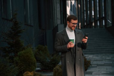 Bir bardak içki ve akıllı telefonla. Resmi kıyafetli yakışıklı dışarıda, iş binasının yanında..