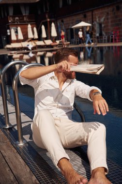 Parlak güneş ışığından kendini gazeteyle kaplıyor. Resmi giyinmiş yakışıklı adam havuzda..