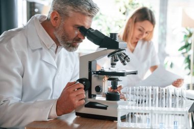 Adam oturuyor ve mikroskop kullanıyor. İki klinik çalışanı içeride birlikte..