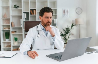 Masanın başında oturuyor. Resmi kıyafetli doktor dolapta çalışıyor..