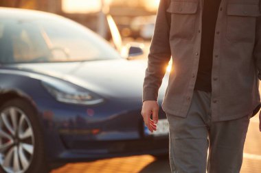 Parlak güneş ışığı. Adam dışarıda elektrikli arabasının yanında duruyor..