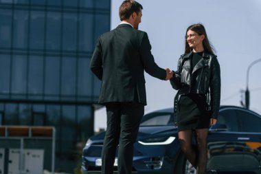 İşadamı ayakta duruyor ve elektrikli arabanın yanında bir kadınla tokalaşıyor..