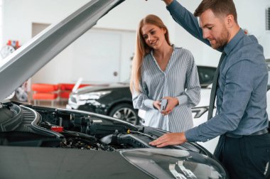 Motor kaputun altında görünüyor. Resmi elbiseli adam elektrikli arabalı kadın müşteriyle birlikte..
