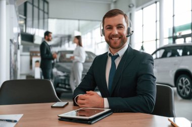 Kulaklıklı adam masanın yanında oturuyor. Araba galerisinde üç kişi birlikte çalışıyor..