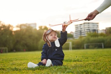 Havacı gözlüklü küçük çocuk sahada oturuyor. Kadın eli ona oyuncak uçak veriyor..
