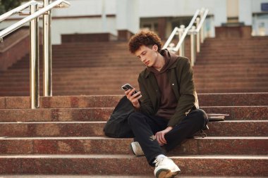 Akıllı telefon ellerdedir. Kıvırcık saçlı genç öğrenci üniversiteye yakın bir yerde..