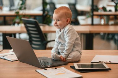 Dizüstü bilgisayarın yanında masada oturuyor. Küçük tatlı erkek bebek modern ofiste..