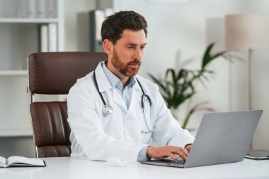 Bilgisayarlı masanın yanında. Resmi kıyafetli doktor dolapta çalışıyor..