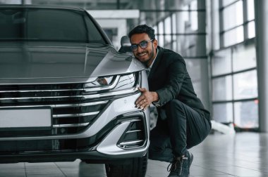 Ulaşımı kucaklıyorum. Takım elbiseli yakışıklı Hintli adam araba galerisinde araçla birlikte..