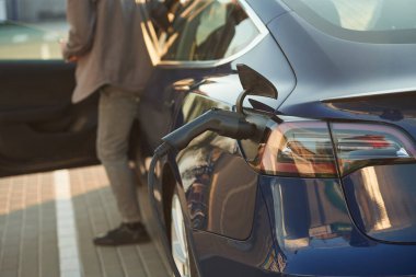 Şarj olan otomobilin arka kısmı. Adam dışarıda elektrikli arabasının yanında duruyor..