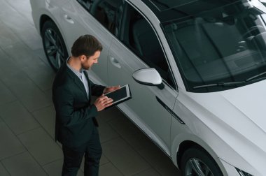 Tabletle en iyi görüntü. Siyah takım elbiseli adam otomobilin yanındaki araba galerisinde..