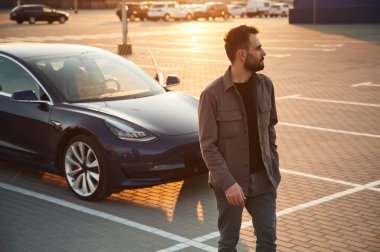 Yakışıklı adam dışarıda elektrikli arabasının yanında duruyor..