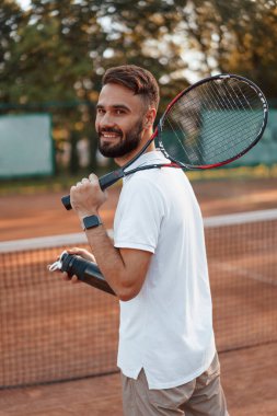 Su ve raketle şişeyi tutuyor. Genç adam güneşli bir günde tenis kortunda..