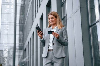 Kredi kartı ve akıllı telefon tutuyor. Resmi giysili kadın iş binasının yakınında dışarıda..