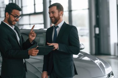Yeni plan. Grafik tabletli otomobilin yanında duruyor. İki işadamı araba galerisinde birlikte çalışıyorlar..