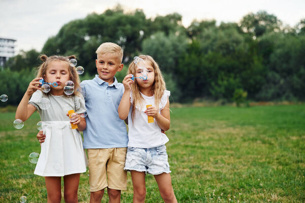 Boy with two girls, playing with bubbles. Kids are having fun on the field at daytime together.