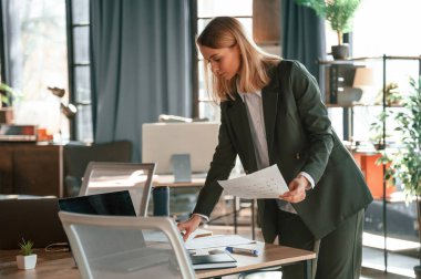 Masanın yanında duruyor. Resmi giyinmiş genç bir bayan ofiste çalışıyor..