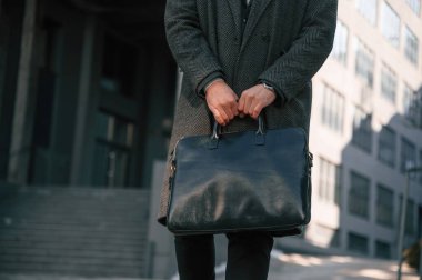 Siyah giysili adam dışarıda duruyor ve çantasını tutuyor..