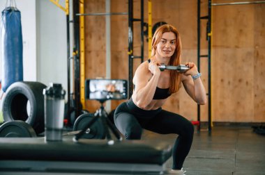 Çömeliyor, akıllı telefondan video çekiyor. Güzel, güçlü kadın spor salonunda..