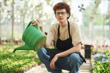 Yeşil sulama kutusu ellerinde. Kıvırcık saçlı ve gözlüklü genç adam serada..