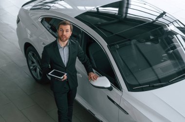 Tabletle en iyi görüntü. Siyah takım elbiseli adam otomobilin yanındaki araba galerisinde..