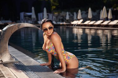 Ayakta, yüzeye yaslanarak. Havuzda mayo giymiş güzel bir kadın var..