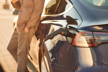 Parlak güneş ışığı. Adam dışarıda elektrikli arabasının yanında duruyor..