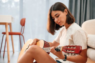 Elinde klasik gitarla. Günlük giysiler içinde güzel bir kadın evde, yurt odasında..